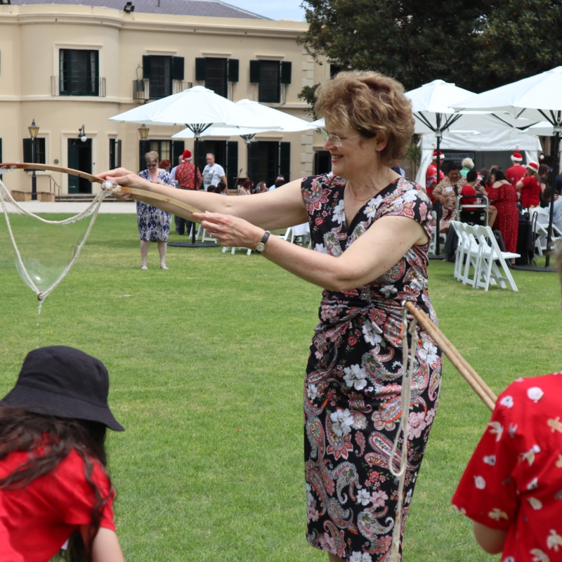Her Excellency using two sticks to blow bubbles at the Legacy Family Christmas Party.